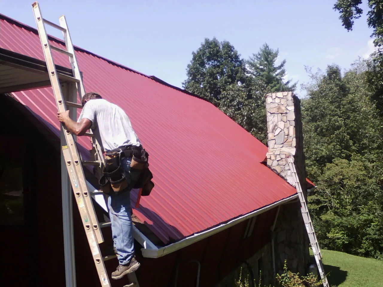 Roofer on a ladder beside a red metal roof and stone chimney — Soffit & Fascia Repair by Roofing Brothers in Lewis